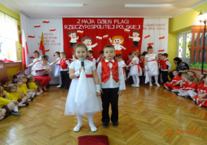 Dziewczynka w białej sukience z czerwonym paskiem i chłopiec w białej koszuli, czerwonej kamizelce i czarnych spodniach recytują wiersz.